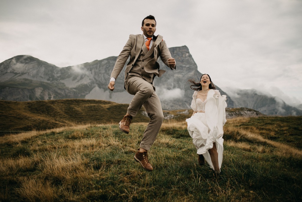 Ein ausgelassenes Bild eines Hochzeitspaares in der Schweiz, im Hintergrund Nebel und Berge, der Bräutigam springt mit Anlauf in die Luft, die Braut lacht aus vollem Herzen