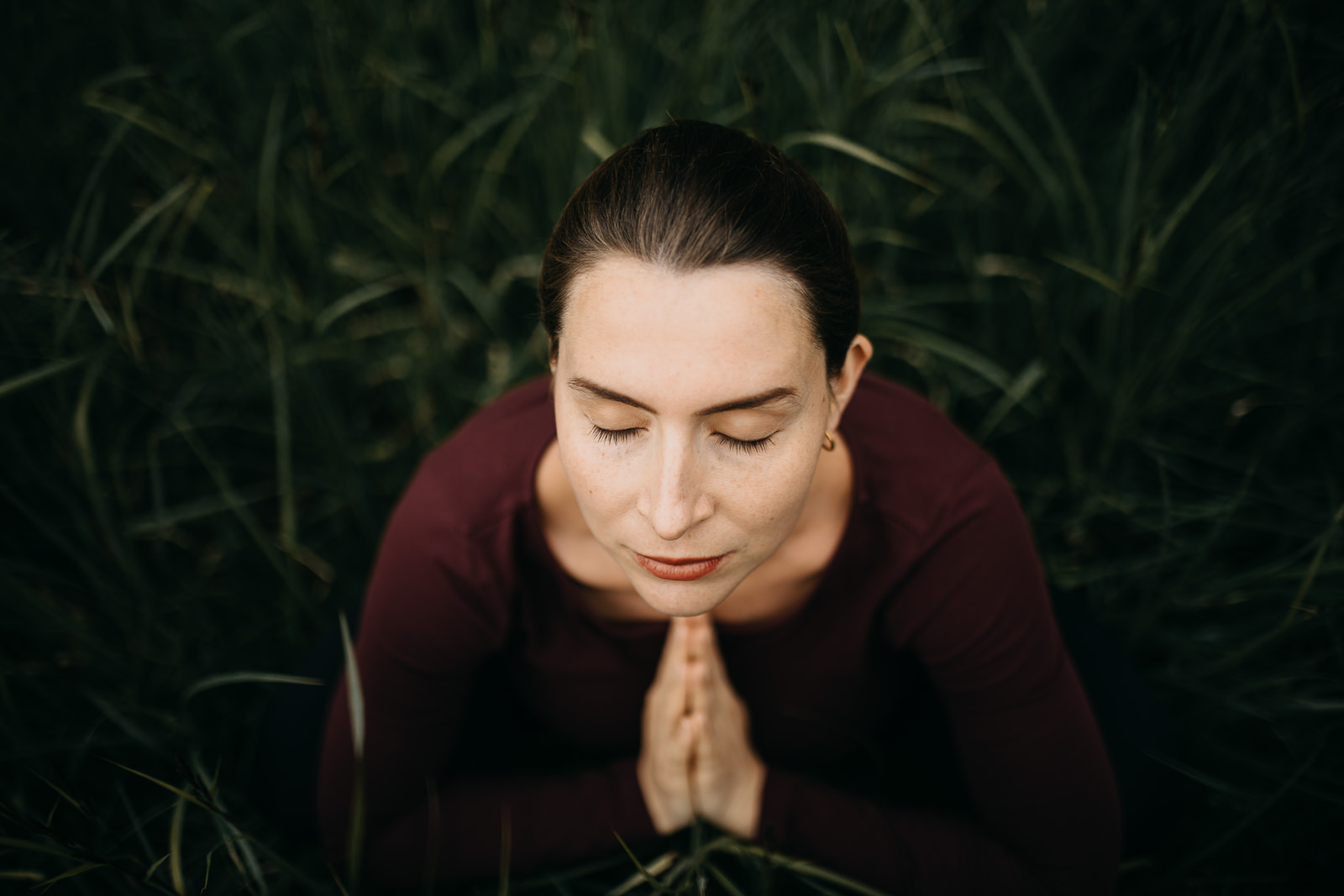 Portrait von Elisa Körperfefühl mit geschlossenen Augen und aufeinandergelegten Händen im Gras sitzend für ein Yogashooting