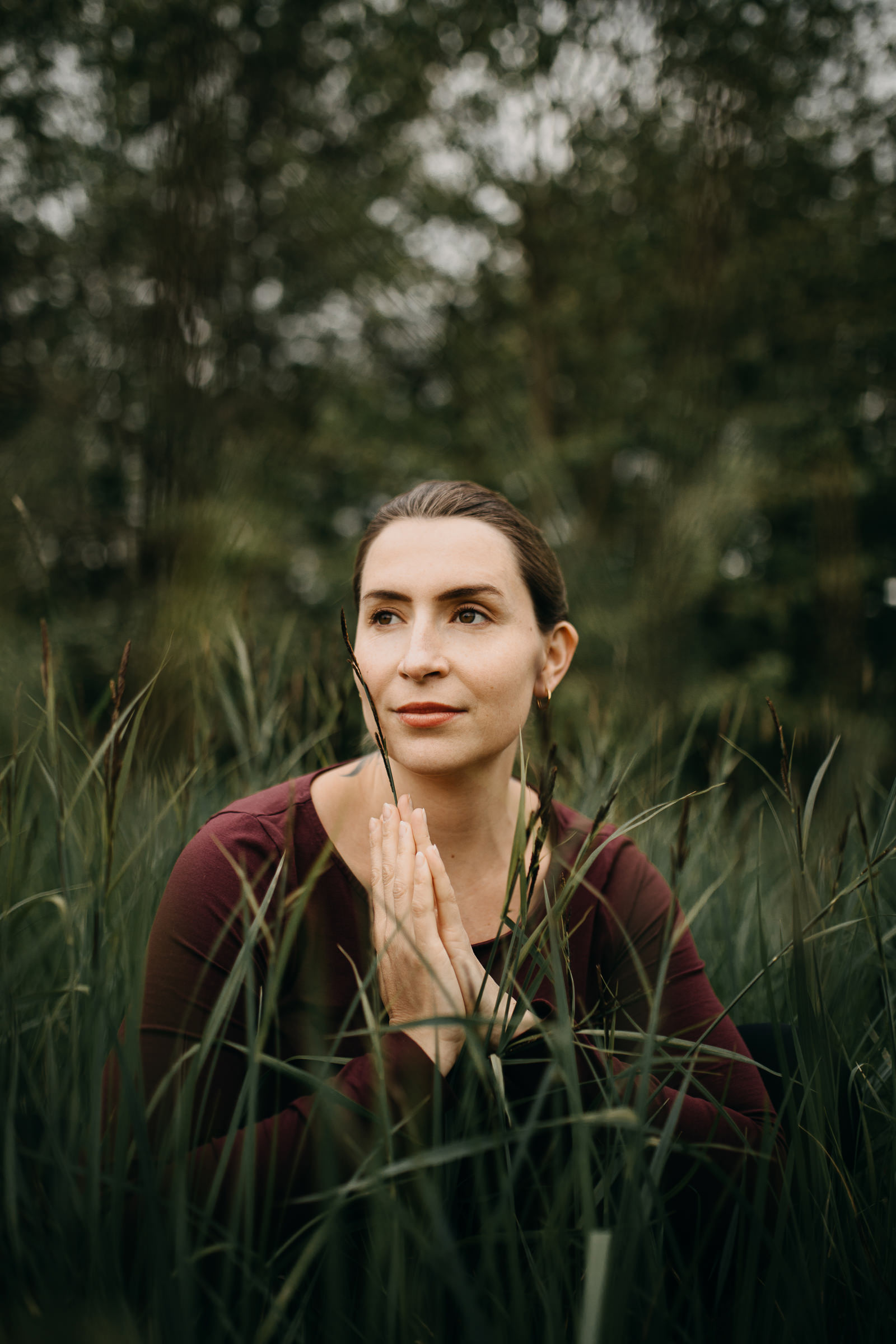 Yogaportrait von Elisa von Körpergefühl im Gras sitzend, mit den Händen aufeinandergelegt und im hohen Gras stehend